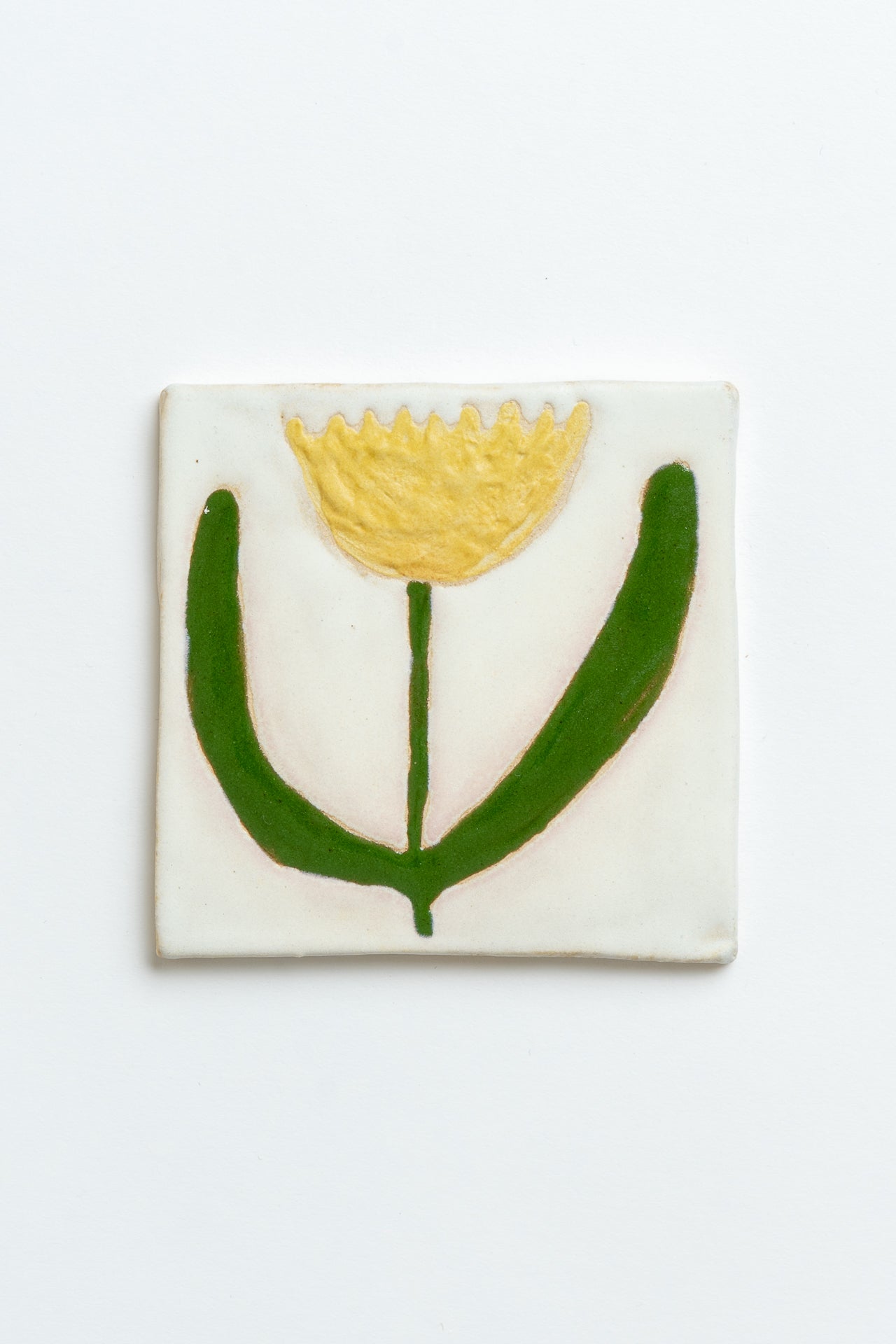 Square ceramic tile with a yellow flower and green leaves on a white background handmade by contemporary British ceramicist Studio Alice Johnson in collaboration with MIMMO Studios
