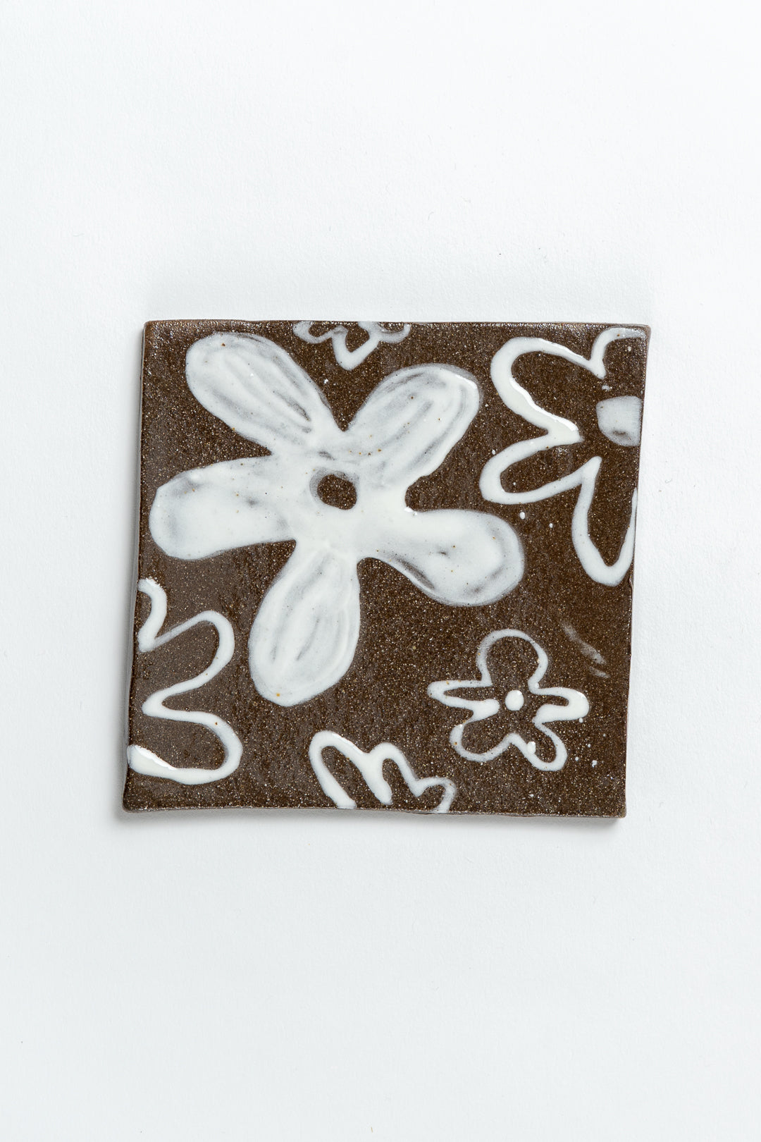 A square ceramic tile with a hand painted brown background and white floral patterns. MIMMO Studios x Studio Alice Johnson Collaboration