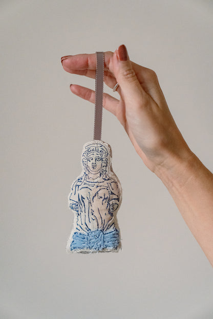 Hand holding a small embroidered figurine against a plain background. Decorative Christmas decoration shaped like a classical figure on a white background, inspired by the caryatid stone ladies in Montpellier Cheltenham. Iconic landmark. MIMMO Studios x Ludio Christmas collaboration