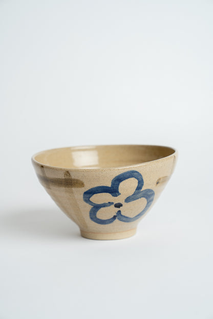 A handmade ceramic bowl with a beige stoneware finish, featuring a hand-painted blue flower motif on the side, set against a plain light gray background.