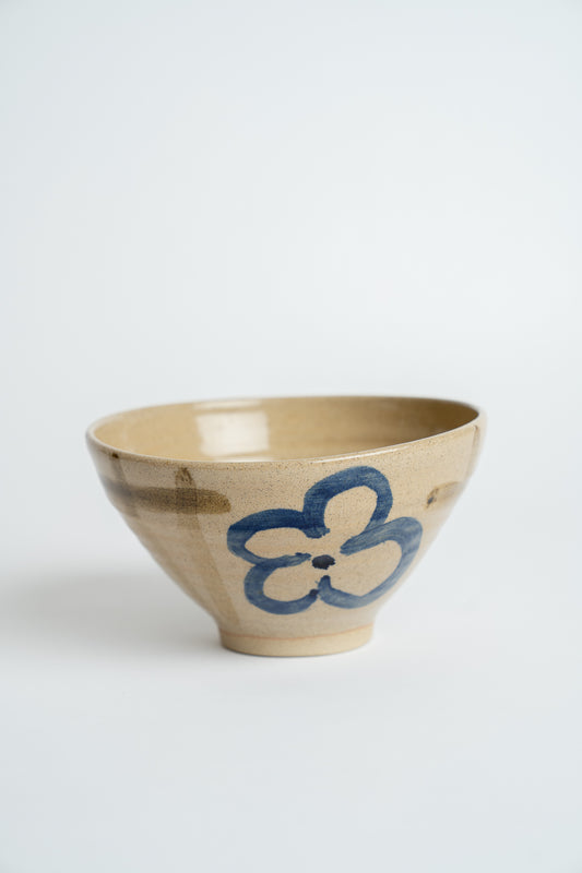 A handmade ceramic bowl with a beige stoneware finish, featuring a hand-painted blue flower motif on the side, set against a plain light gray background.